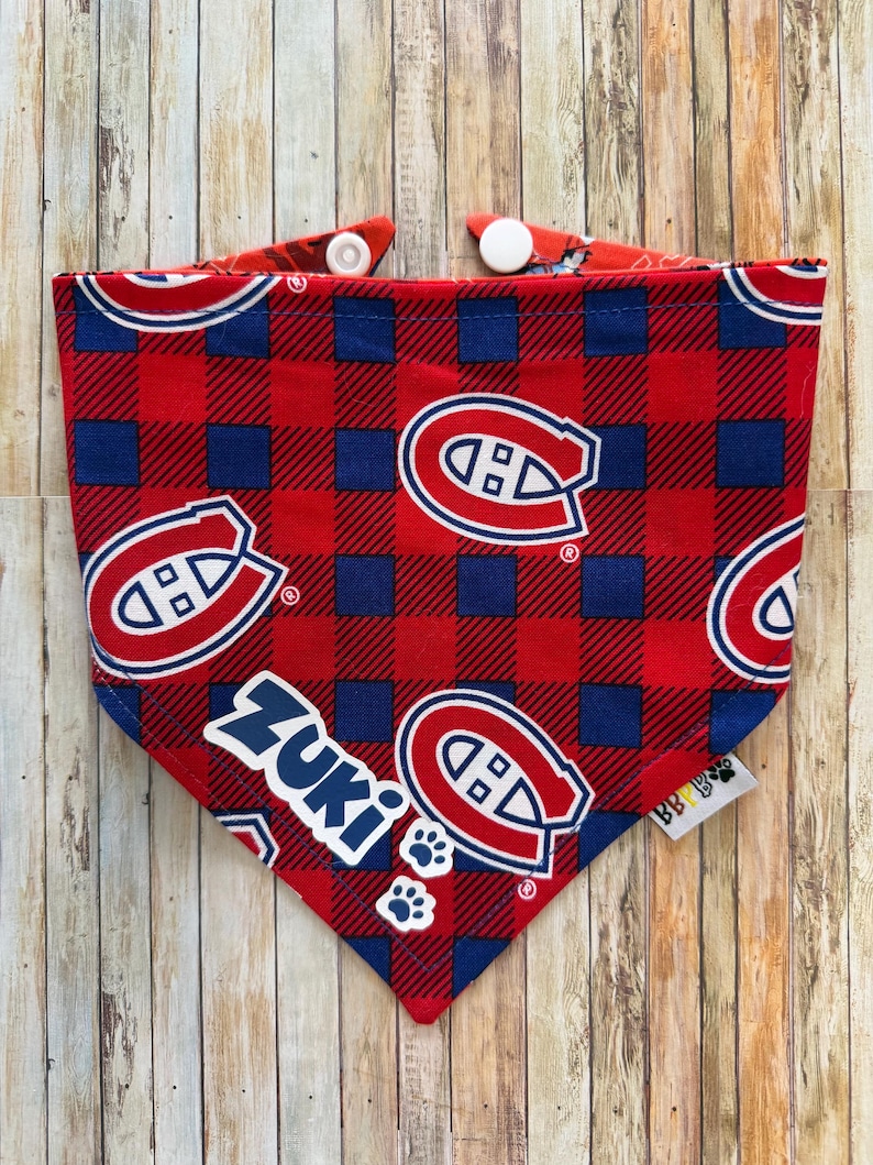 May include: Red, blue, and white plaid dog bandana with the text "Zuki" and paw prints. The bandana features the logo of the Montreal Canadiens hockey team. The bandana has white snaps for easy closure.