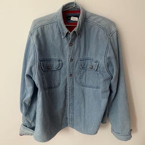 May include: Light blue denim shirt with two chest pockets and button fastenings. The shirt is hanging on a metal hanger. The collar has a red lining. A classic style denim shirt.