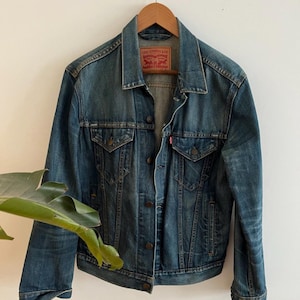 May include: A blue denim jacket with a classic design, featuring two chest pockets and button closures. The jacket is hanging on a wooden hanger against a white wall. A red and white label is visible on the jacket.