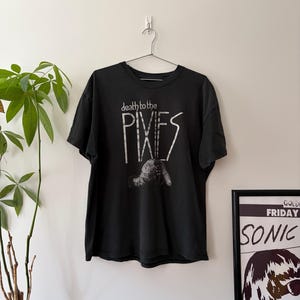 May include: A black short-sleeve t-shirt featuring the white text "death to the PIXIES" printed on the front. Below the text is a white graphic illustration of a figure. The shirt is displayed on a metal hanger against a plain wall.