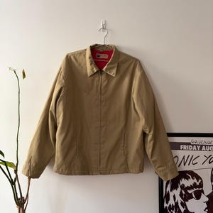May include: A tan zip-up jacket with a red interior lining hangs on a metal hanger against a white wall. The jacket has a collared neckline and two front pockets. A poster with black and white graphics is visible on the right.