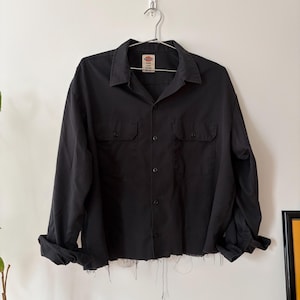 May include: Black long-sleeve shirt with two chest pockets and a frayed hem. The shirt is hanging on a metal hanger against a white wall. The shirt has a button-down front and a collar.