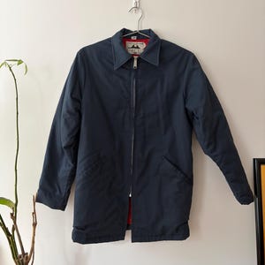 May include: A navy blue jacket with a full-length zipper, a collar, and two front pockets. The jacket has a red lining and is hanging on a metal hanger. The jacket is a medium length.