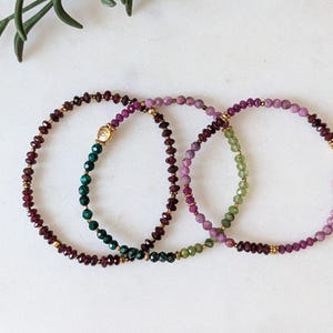 May include: Two delicate beaded bracelets featuring faceted gemstone beads in shades of deep red, magenta, and green. The bracelets are accented with small gold-toned metal spacers and a single gold-toned charm on one of the strands.