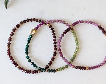 Könnte beinhalten: Zwei zarte Perlenarmbänder mit facettierten Edelsteinperlen in Dunkelrot, Magenta und Grün. Die Armbänder sind mit kleinen goldfarbenen Metallzwischenstücken und einem einzelnen goldfarbenen Anhänger an einem der Stränge verziert.