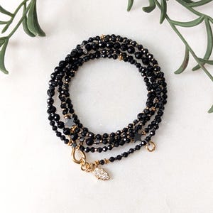 Könnte beinhalten: Ein mehrsträngiges Armband mit kleinen, facettierten schwarzen Perlen, goldfarbenen Akzenten und zwei dunklen sternförmigen Anhängern. Ein goldfarbener Karabinerverschluss sichert das Schmuckstück, das einen kleinen Hamsa-Hand-Anhänger enthält.