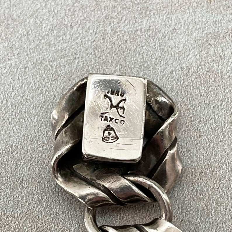 K&ouml;nnte beinhalten: Nahaufnahme eines silbernen Armbands mit einem gedrehten Design. Das Armband hat ein rechteckiges Schild mit dem Text "Taxco" und einem Symbol.