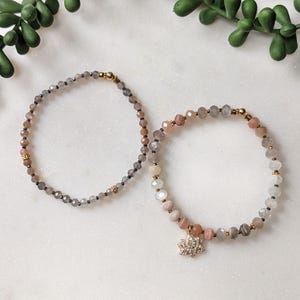 May include: Two beaded bracelets. One is primarily gray with gold accents. The other is a mix of pink, gray, and white beads, with a gold charm featuring a flower design. Both bracelets are on a white marble surface.