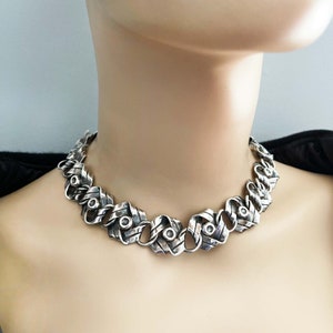 K&ouml;nnte beinhalten: Eine silberne Halskette mit einem geflochtenen Kettendesign. Die Kette besteht aus dickem, strukturiertem Silber und hat eine Reihe von runden Silberakzenten.