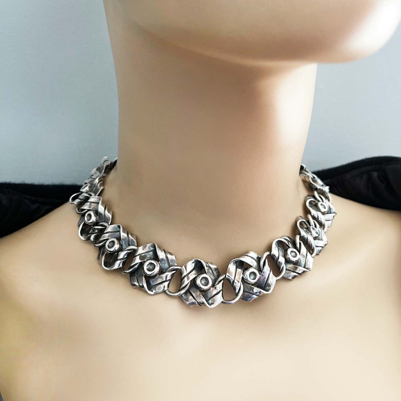 K&ouml;nnte beinhalten: Eine silberne Halskette mit einem geflochtenen Kettendesign. Die Kette besteht aus dickem, strukturiertem Silber und hat eine Reihe von runden Silberakzenten.
