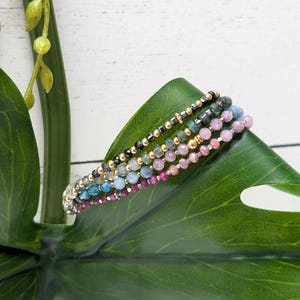 May include: A collection of beaded bracelets in various colors, including pink, blue, green, and gold. The bracelets are made with small, round beads and are displayed on a large green leaf. The background is a white wooden surface.