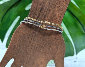 Könnte beinhalten: Ein Stapel von drei Armbändern mit Perlen in Gold, Silber und Dunkelgrau. Die Armbänder werden auf einem handförmigen Schmuckhalter aus Holz vor einem Hintergrund aus grünen Blättern präsentiert.