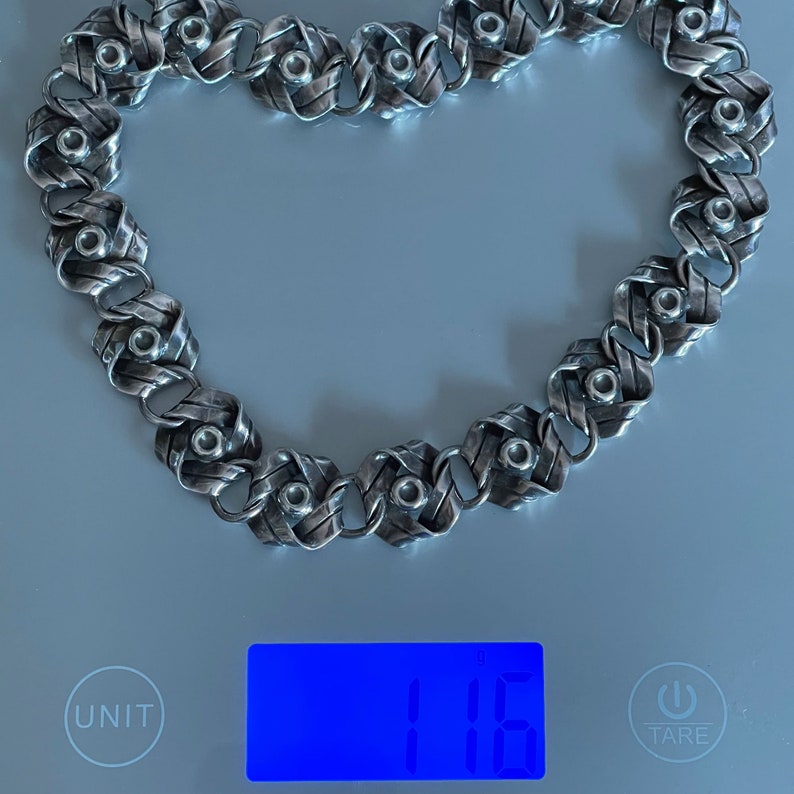 K&ouml;nnte beinhalten: Eine silberne Kettenkette mit einem einzigartigen gewebten Design und kleinen runden Akzenten. Die Kette liegt auf einer digitalen Waage, die 11,6 Gramm anzeigt.