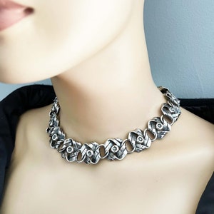 K&ouml;nnte beinhalten: Eine silberne Choker-Kette mit geflochtenem Design und runden Akzenten. Die Kette ist aus Metall und hat eine dicke, strukturierte Optik.