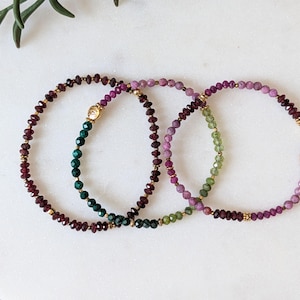 May include: Three beaded bracelets in shades of burgundy, purple, green, and gold. The bracelets are made of small, faceted beads strung together. The bracelets are arranged in a curved shape, with a gold clasp on one of the bracelets.