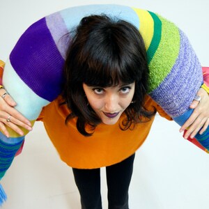 May include: A person wearing a bright orange sweater and black pants is holding a long, colorful, striped knitted pillow over their shoulders.