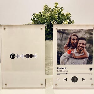 Pu&ograve; includere: Due cornici fotografiche in acrilico trasparente. Una presenta una foto di una coppia con i dettagli della canzone "Perfect" di Ed Sheeran. L'altra cornice ha un codice Spotify. Entrambe sono esposte su una superficie bianca.