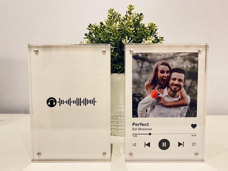 Pu&ograve; includere: Due cornici fotografiche in acrilico trasparente. Una presenta una foto di una coppia con i dettagli della canzone "Perfect" di Ed Sheeran. L'altra cornice ha un codice Spotify. Entrambe sono esposte su una superficie bianca.