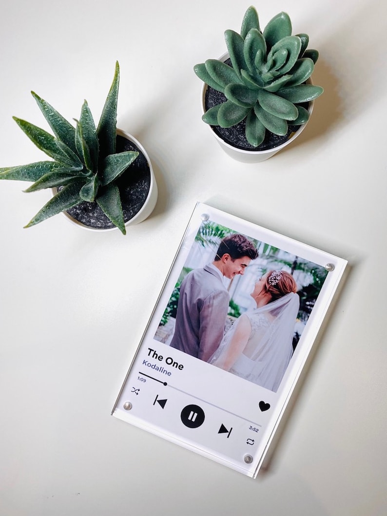 Pu&ograve; includere: Un blocco fotografico in acrilico trasparente con una foto di matrimonio, il testo "The One" e un'interfaccia del lettore musicale. Due piccole piante grasse in vaso sono sulla superficie bianca. Il blocco fotografico &egrave; tenuto insieme da viti argentate.