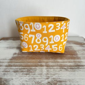 May include: A small, rectangular fabric basket with a bright yellow exterior and interior. The exterior is patterned with white numbers 1 through 10, with red stitching details. The basket sits on a wooden surface.