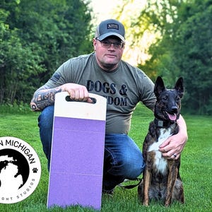 May include: A man wearing a green shirt with the text "Dogs & Freedom" is kneeling on the grass with a dog. He is holding a purple training pad with a white handle. The training pad has a "Made in Michigan" logo with a paw print and the text "2 Dog Night Studios".