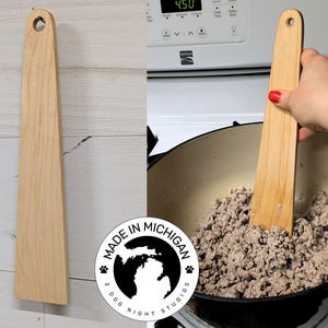 May include: A wooden spatula with a rounded handle and a wide, flat blade. The spatula is made in Michigan and is being used to stir ground meat in a black pot.