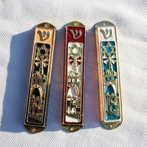 Pode incluir: Três mezuzá com detalhes dourados e fundos de cores diferentes. As mezuzá têm uma estrela de David, uma letra hebraica e uma representação do Muro das Lamentações.