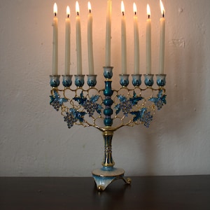 May include: A lit menorah with nine candles. The candle holders are blue and gold, with a decorative grape vine design. The menorah is placed on a dark brown surface against a white wall.