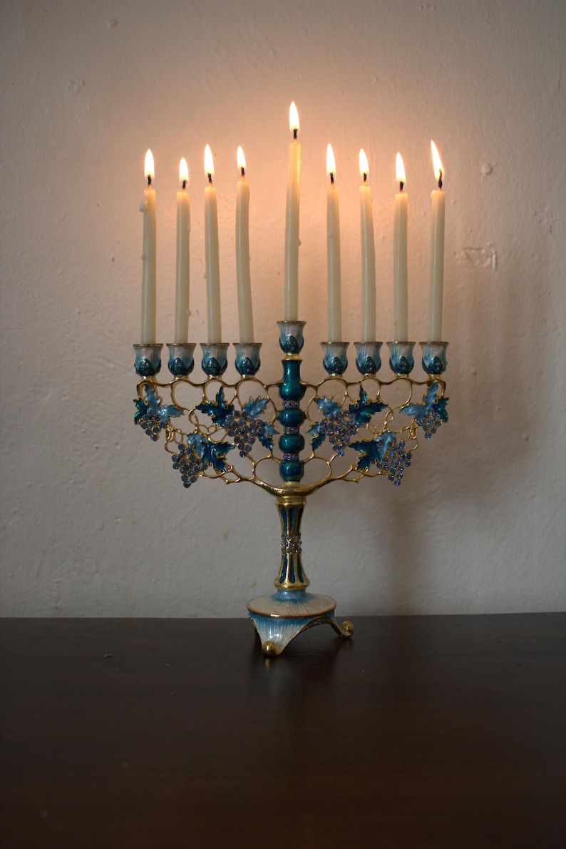 May include: A lit menorah with nine candles. The candle holders are blue and gold, with a decorative grape vine design. The menorah is placed on a dark brown surface against a white wall.