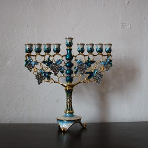 May include: A teal and gold menorah with nine candle holders. The design includes grape vines and leaves. The base and candle holders are teal, with gold accents. The menorah is a decorative item.