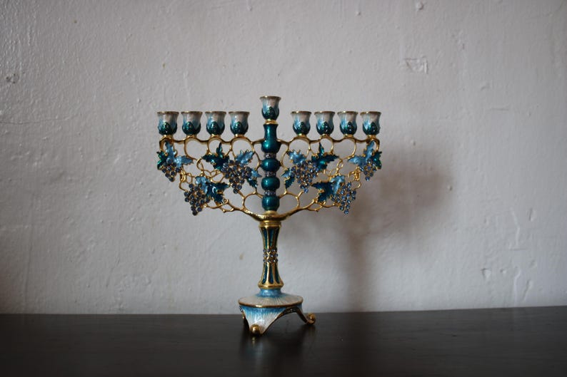 May include: A teal and gold menorah with nine candle holders. The design includes grape vines and leaves. The base and candle holders are teal, with gold accents. The menorah is a decorative item.