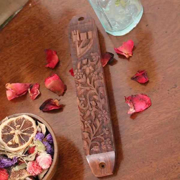 May include: A wooden mezuzah with a carved floral design and the Hebrew letter "Shin" on a brown wooden surface with rose petals scattered around it.