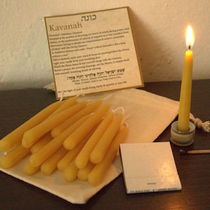 Könnte beinhalten: Ein Set mit 20 gelben Bienenwachskerzen, eine kleine weiße Tasche, eine weiße Karte mit dem Text "Kavanah" und hebräischem Text, ein kleiner weißer Notizblock, eine brennende gelbe Kerze in einem silbernen Halter und ein Streichholz auf einer Holzoberfläche.