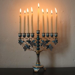May include: A lit menorah with nine candles. The candle holders are teal and gold, with a grape vine design. The candles are lit, with flames at the top. The menorah is on a dark surface, with a white wall in the background.
