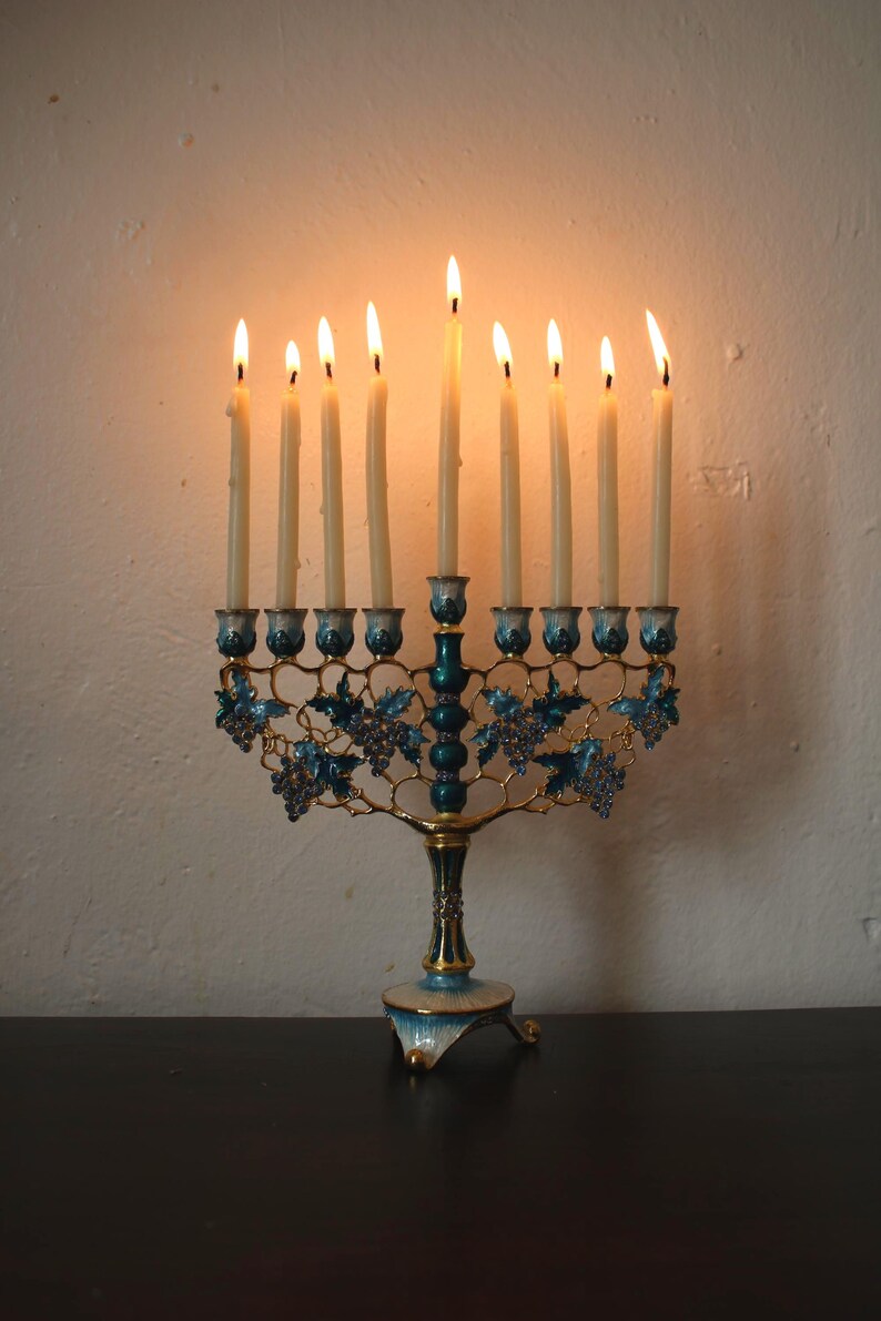 May include: A lit menorah with nine candles. The candle holders are teal and gold, with a grape vine design. The candles are lit, with flames at the top. The menorah is on a dark surface, with a white wall in the background.