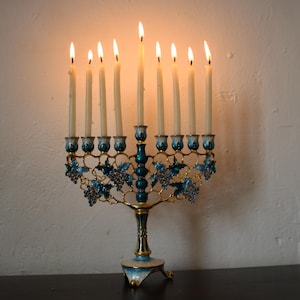 May include: A Hanukkah menorah with nine lit candles. The teal and gold candle holders feature a grape vine design. The candles are alight, with flames visible. The menorah sits on a dark surface against a white wall.