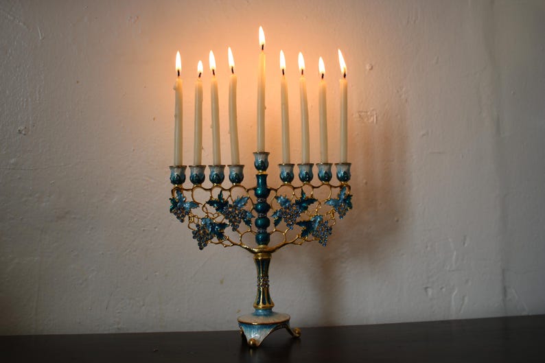 May include: A Hanukkah menorah with nine lit candles. The teal and gold candle holders feature a grape vine design. The candles are alight, with flames visible. The menorah sits on a dark surface against a white wall.