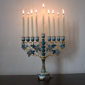 May include: A lit menorah with nine candles. The brass-coloured base is decorated with blue enamel and grape designs. The candles are lit, casting a warm glow. The menorah is placed on a dark wooden surface.