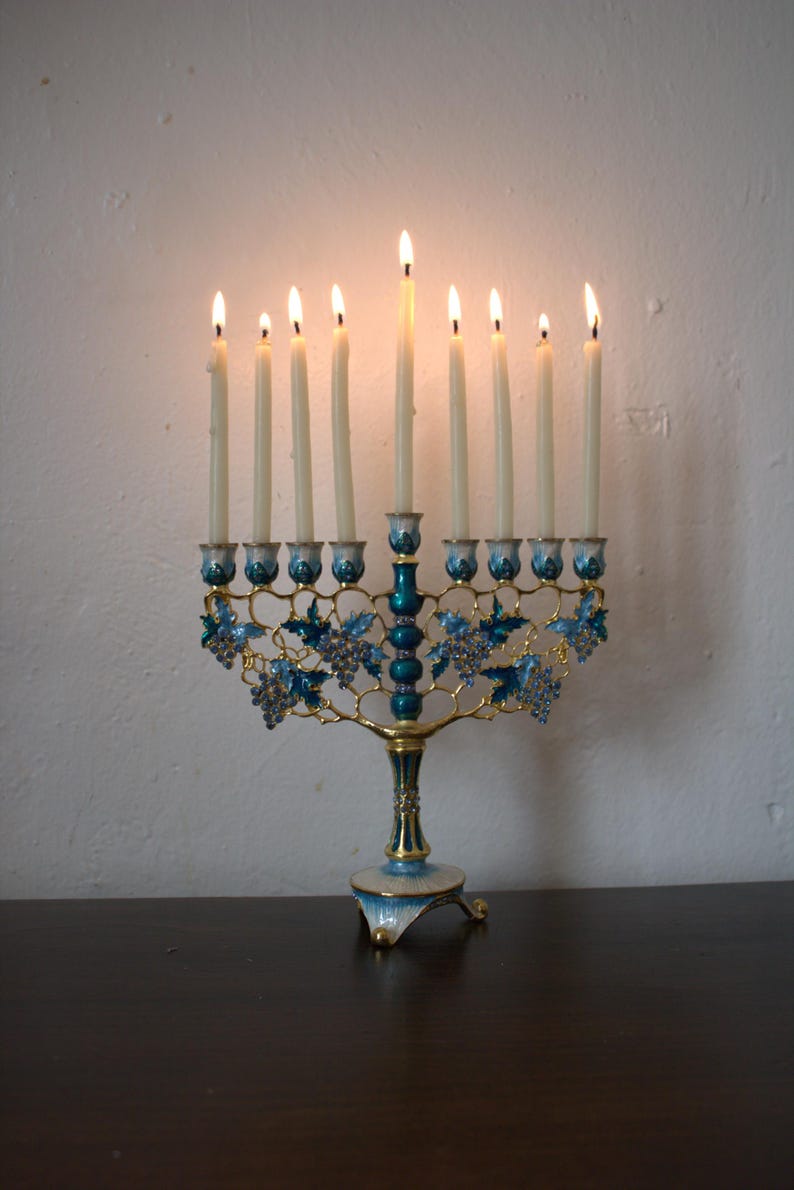 May include: A lit menorah with nine candles. The brass-coloured base is decorated with blue enamel and grape designs. The candles are lit, casting a warm glow. The menorah is placed on a dark wooden surface.