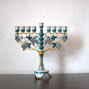 May include: A decorative menorah with a gold-coloured frame and teal accents. The menorah features grape cluster and leaf designs in blue and teal. The base and candle holders are also teal and gold.