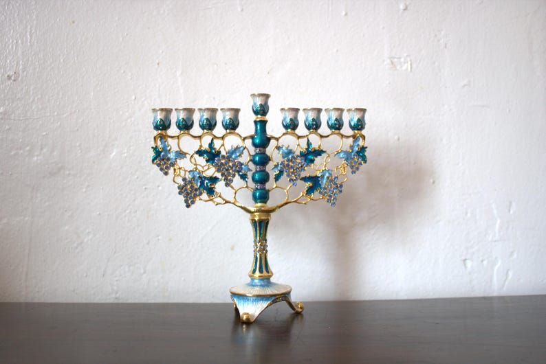 May include: A decorative menorah with a gold-coloured frame and teal accents. The menorah features grape cluster and leaf designs in blue and teal. The base and candle holders are also teal and gold.