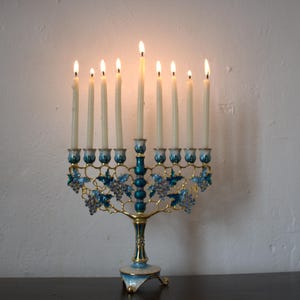May include: A lit menorah with nine candles. The candle holder is gold-coloured with teal and blue accents, including floral and grape vine designs. The candles are lit, with flames visible.