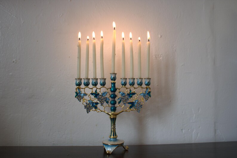 May include: A lit menorah with nine candles. The candle holder is gold-coloured with teal and blue accents, including floral and grape vine designs. The candles are lit, with flames visible.