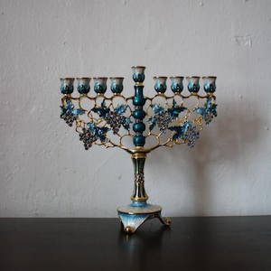 May include: A decorative menorah with nine candle holders. The menorah has a gold-coloured frame with blue and teal enamel accents. Blue grape clusters and leaves adorn the arms. The base is white and blue with gold trim.