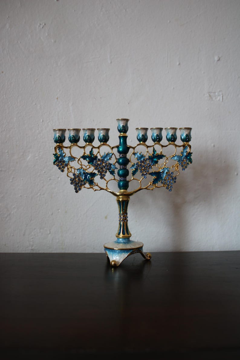May include: A decorative menorah with nine candle holders. The menorah has a gold-coloured frame with blue and teal enamel accents. Blue grape clusters and leaves adorn the arms. The base is white and blue with gold trim.
