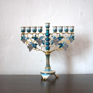 May include: A decorative menorah with a teal and gold finish. The candle holders are white and teal. The menorah features blue grape and leaf accents. The base is white and teal with gold trim.