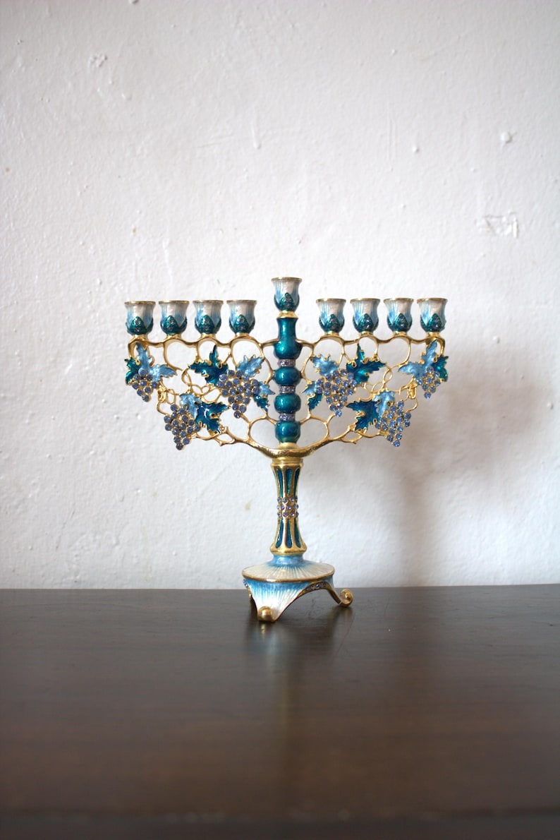 May include: A decorative menorah with a teal and gold finish. The candle holders are white and teal. The menorah features blue grape and leaf accents. The base is white and teal with gold trim.