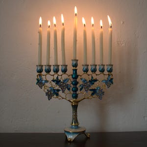 May include: A lit menorah with nine candles. The candle holders are teal and gold, with blue grape vine designs. The candles are lit, casting a warm glow. The menorah sits on a dark surface against a white wall.