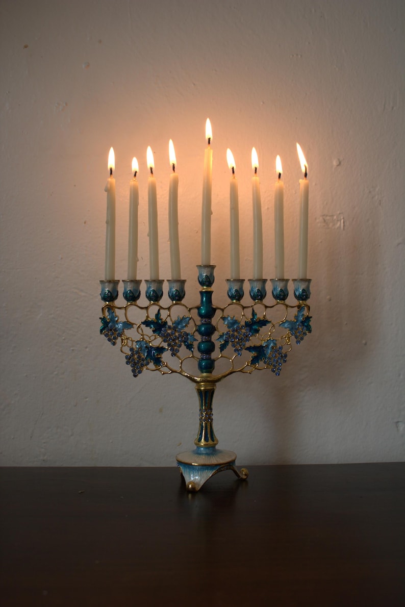 May include: A lit menorah with nine candles. The candle holders are teal and gold, with blue grape vine designs. The candles are lit, casting a warm glow. The menorah sits on a dark surface against a white wall.
