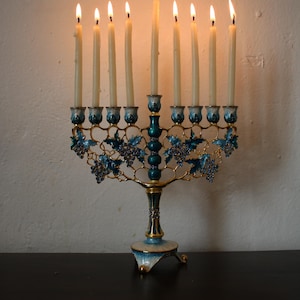 May include: A lit menorah with nine candles. The candle holders are teal and gold, with blue grape vine designs. The base is teal and gold. The candles are lit, with flames at the top.